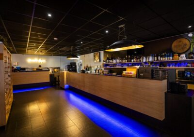 De bar van Yumi Sushi & Grill heeft een strak en eigentijds design met een lange houten counter en sfeervolle blauwe LED-verlichting. Achter de bar staan stijlvolle rekken met een uitgebreide selectie dranken en glazen.