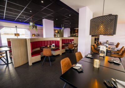 Interieur van Yumi Sushi & Grill met moderne zitplaatsen, warme verlichting en stijlvolle details zoals hangende planten en een opvallende lichtarmatuur. De gedekte tafels met chopsticks en zwarte borden benadrukken de verfijnde Aziatische sfeer van het restaurant. De half ronde zithoeken met rode banken bieden extra comfort en privacy, perfect voor een gezellige avond met vrienden of familie.