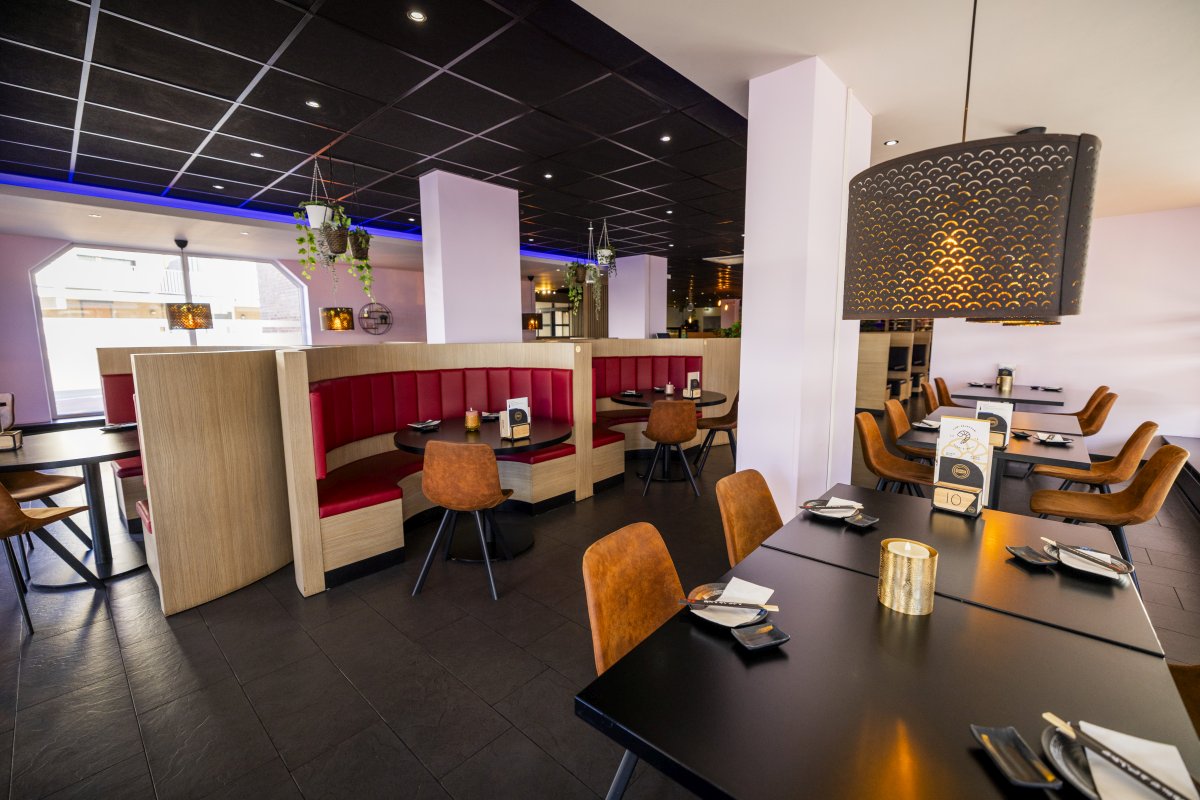 Zithoeken Interieur van Yumi Sushi & Grill met moderne zitplaatsen, warme verlichting en stijlvolle details zoals hangende planten en een opvallende lichtarmatuur. De gedekte tafels met chopsticks en zwarte borden benadrukken de verfijnde Aziatische sfeer van het restaurant. De half ronde zithoeken met rode banken bieden extra comfort en privacy, perfect voor een gezellige avond met vrienden of familie.